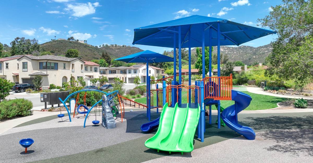 A blue community playground contains three green slides, a climbing wall, two blue slides, and two large umbrellas for shade.