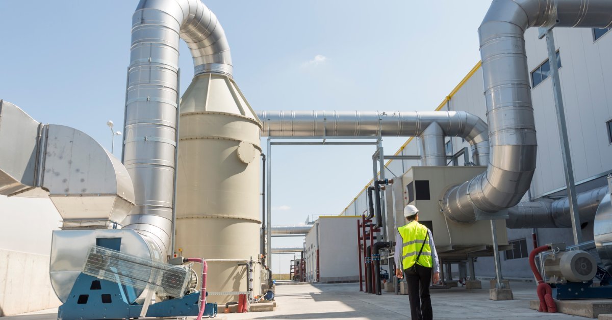 A person wearing a bright yellow safety vest and hard hat walks between oversized metal tubes and tan holding tanks.