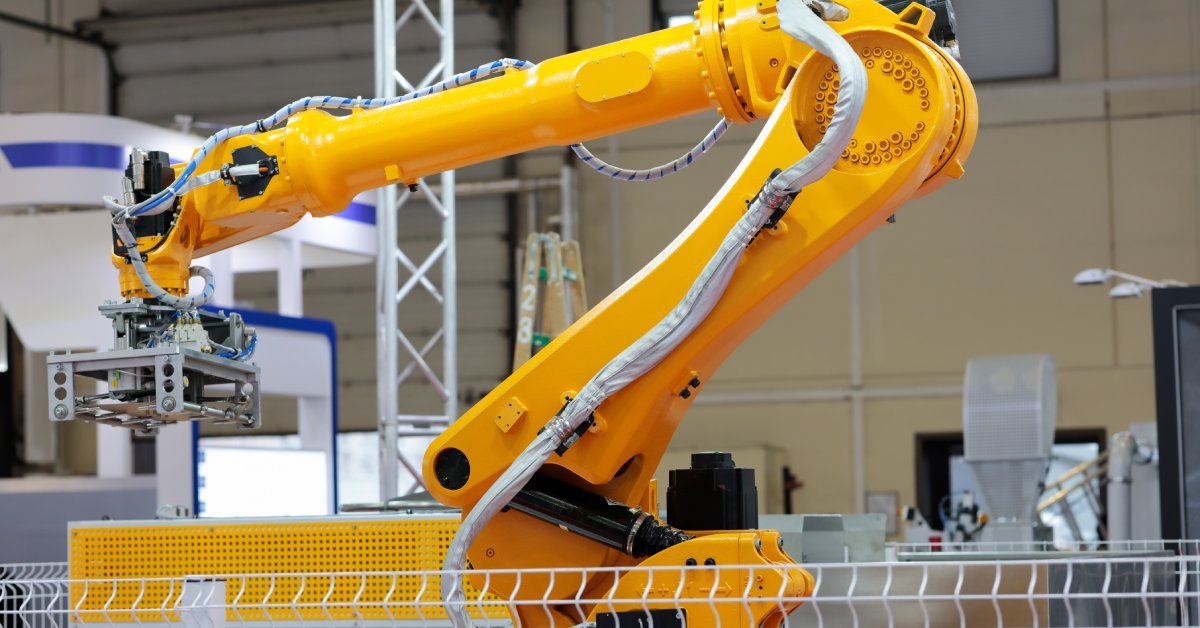 A yellow robotic arm with white wires appears behind a metal fence. Other machines appear behind it.