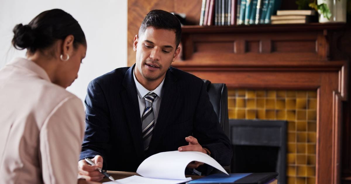 A man dressed in a suit and tie is showing a woman wearing a pink blazer and earrings where to sign a document.