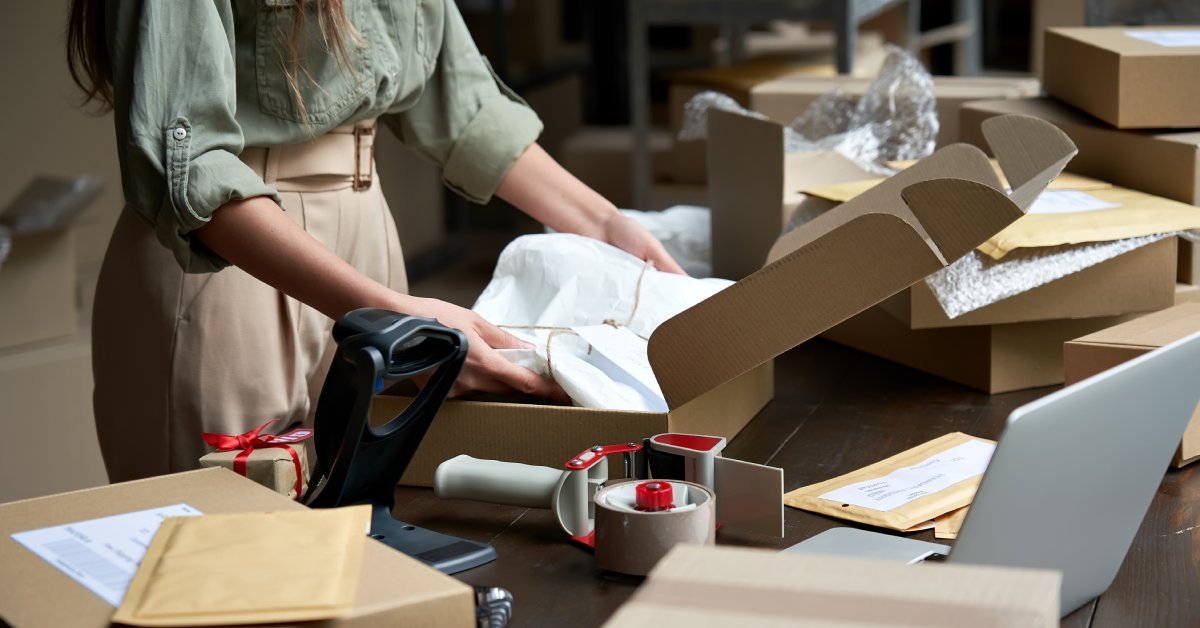 A woman prepares a package to ship. There is a roll of tape, a scanner, and more packing materials nearby.