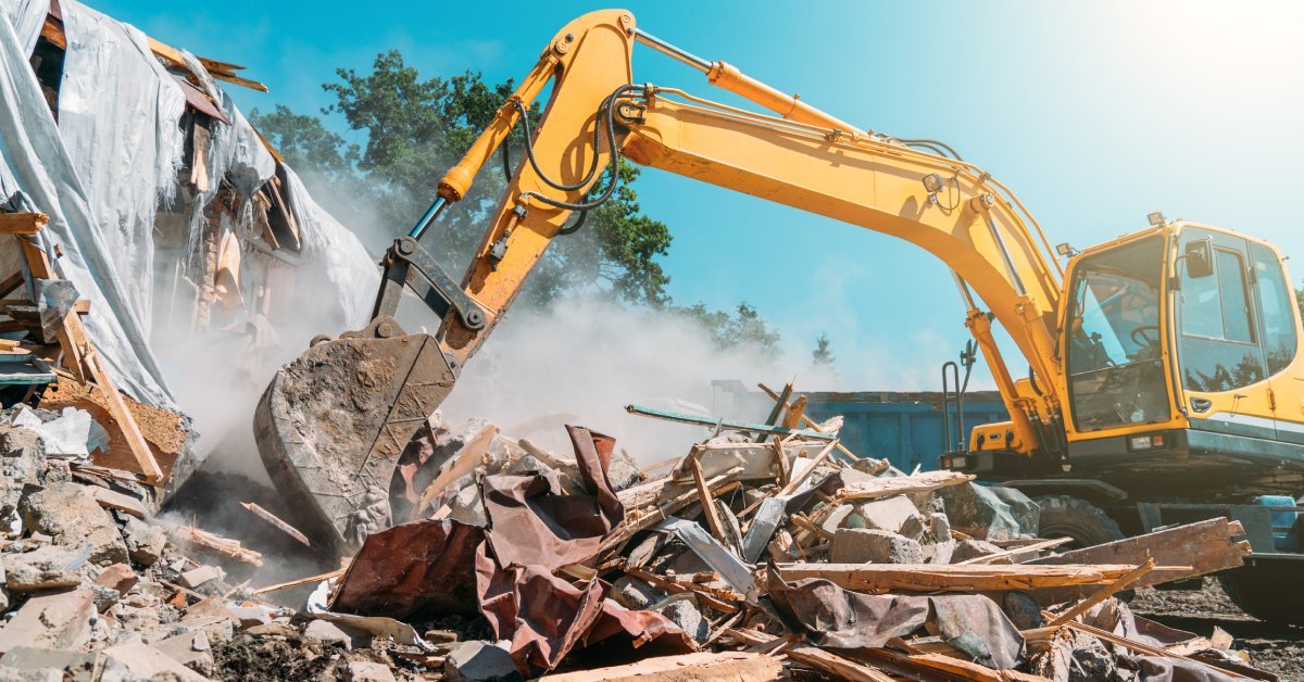 An excavator operator extends the boom and uses the bucket attachment to demolish a building under a sunny blue sky.