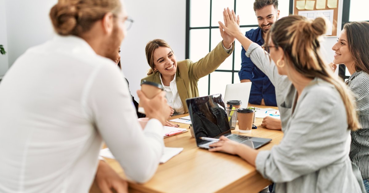 A group of young adults sitting around a wooden table in an office setting. Two of them are smiling and high fiving.