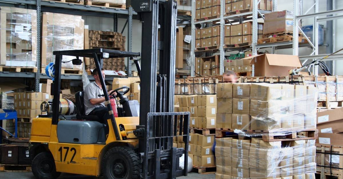 An operator driving a forklift through a warehouse filled with boxes stacked on pallet racking shelves.