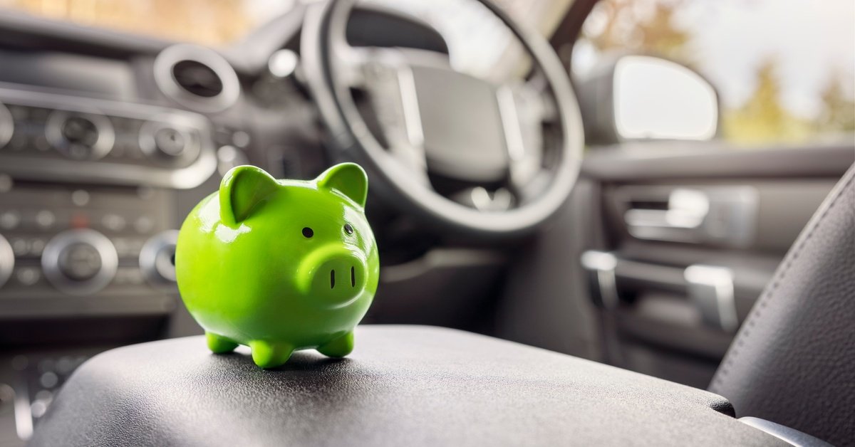 A green piggy bank sits on top of a car's center console armrest. The car's dashboard is blurred in the background.