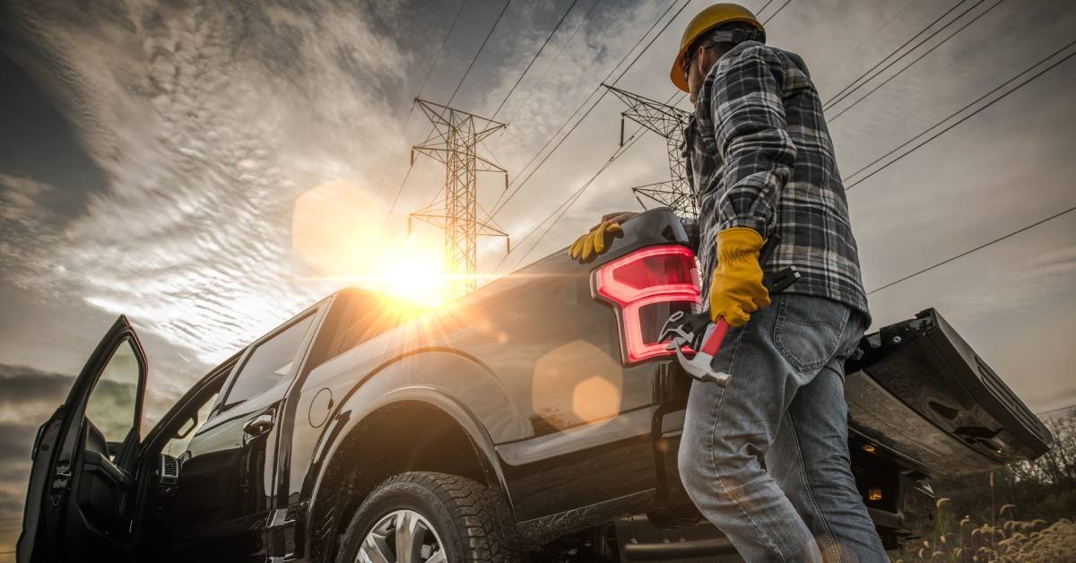 A worker in a hardhat and plaid shirt approaches his truck from behind, holding a hammer, while the sun sets in front.