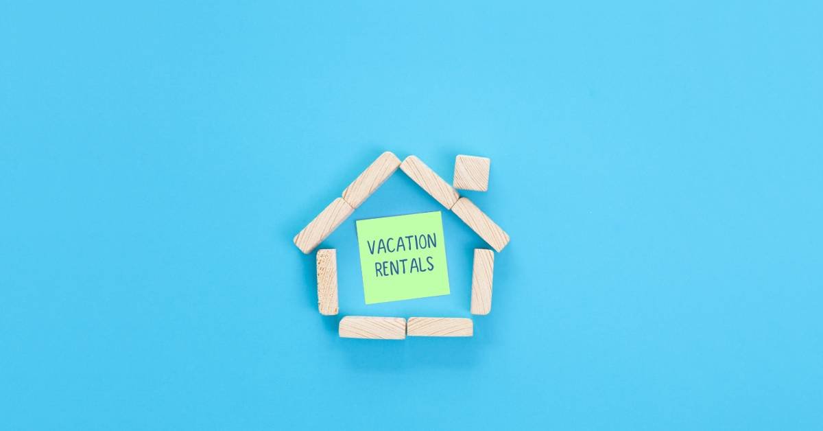 Wooden blocks arranged in the shape of a home with a note inside reading “Vacation Rentals” against a blue background.