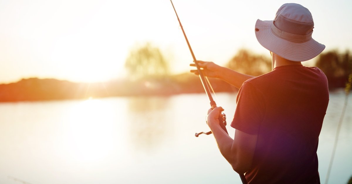 A man wearing a brimmed hat and sunglasses standing on the shore while fishing with the sun shining down on him.