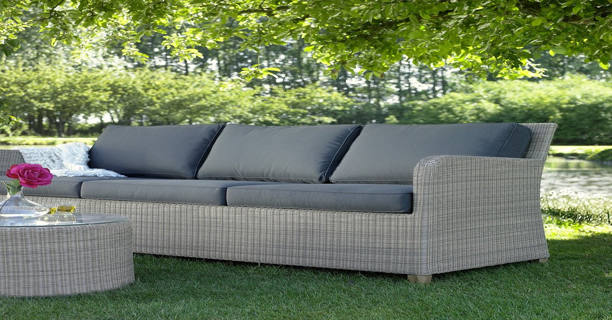 A gray couch with a woven frame and dark-gray cushions outside on a patch of grass with a cylindrical table in front of it.