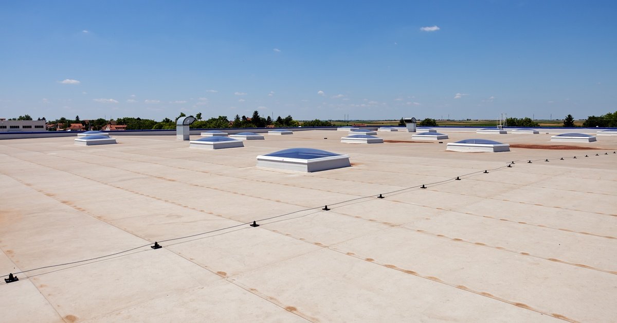 The flat roof of a commercial building on a sunny day. It has a beige membrane and several skylights dotting the surface.
