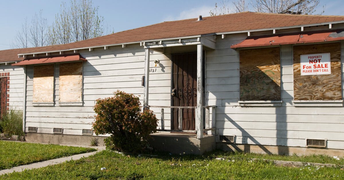 An empty and abandoned home with boarded up windows, faded siding, and a small front porch that's falling apart.