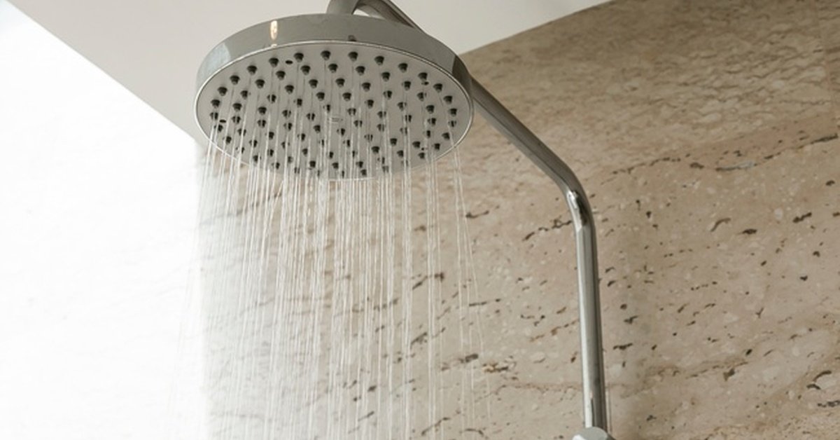 A narrow silver pipe leading up a shower wall to a wide, flat showerhead that is spraying jets of water.