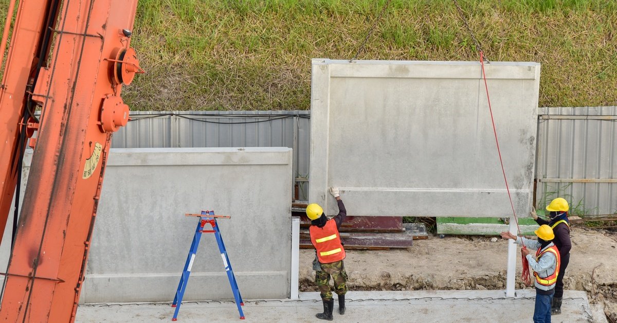 Three construction workers in hard hats and safety vests using a large crane to lift a concrete wall.