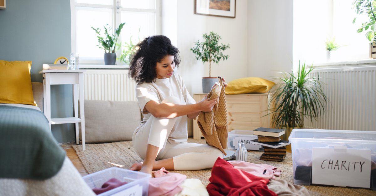 A woman sits on the floor of a bedroom, organizing her clothes and putting some into containers labeled "Charity."