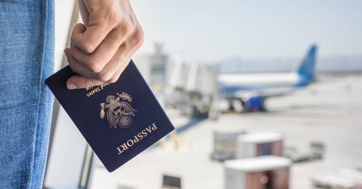 A traveler holding their passport near the airport window as they look out and watch a plane deboard.