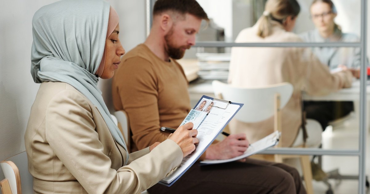 A woman wearing a hijab sits next to a man wearing a brown shirt while writing on a document on a clipboard.