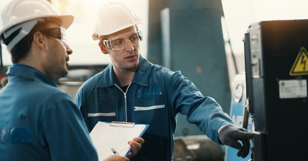 Two manufacturing workers wearing protective gear standing next to a control panel. One man holds a clipboard.