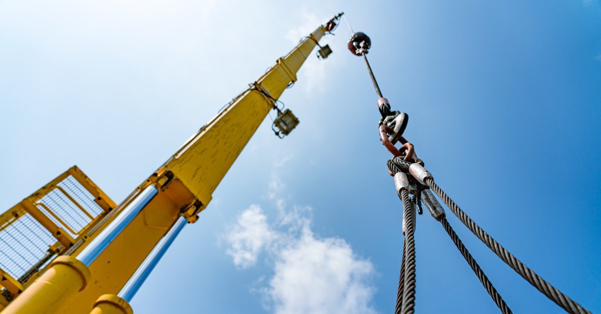 The hydraulic cylinders of an offshore oil rig crane lifting a load held by multiple cable wire slings.