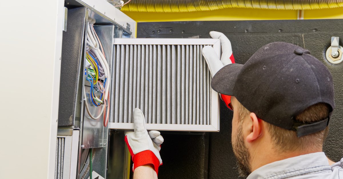 A man in a gray shirt, black hat, and white gloves replaces a dirty air filter in industrial equipment.
