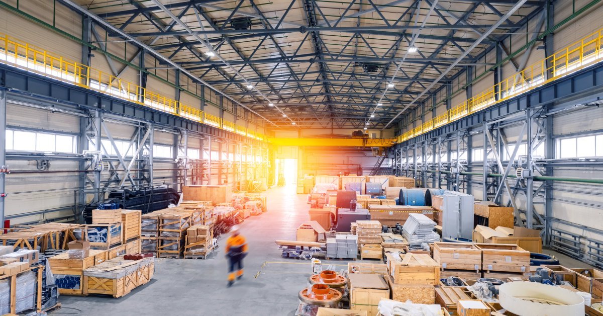 An industrial warehouse that stores materials in wooden boxes. One worker is walking in neon orange safety gear.