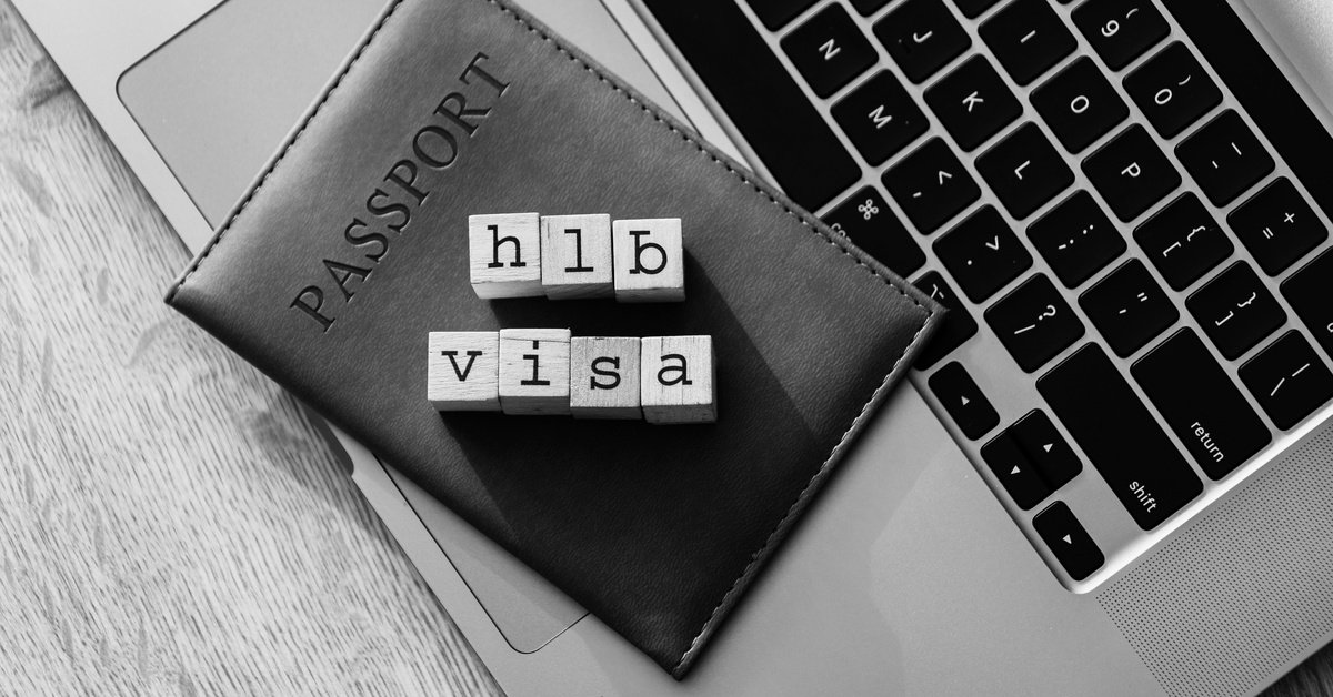 Wooden blocks that spell out "H1B Visa" sits on top of a black passport holder and a laptop on a gray table.