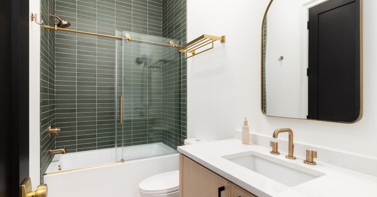 An updated bathroom features a shower with a glass door, green tile, a mirror, and a white sink with gold fixtures.