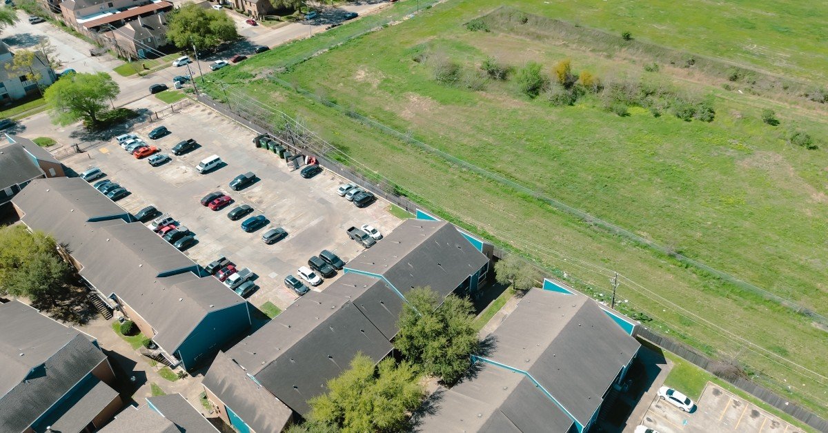 An aerial view of an apartment complex with various buildings and parking lot next to a plot of undeveloped green land.