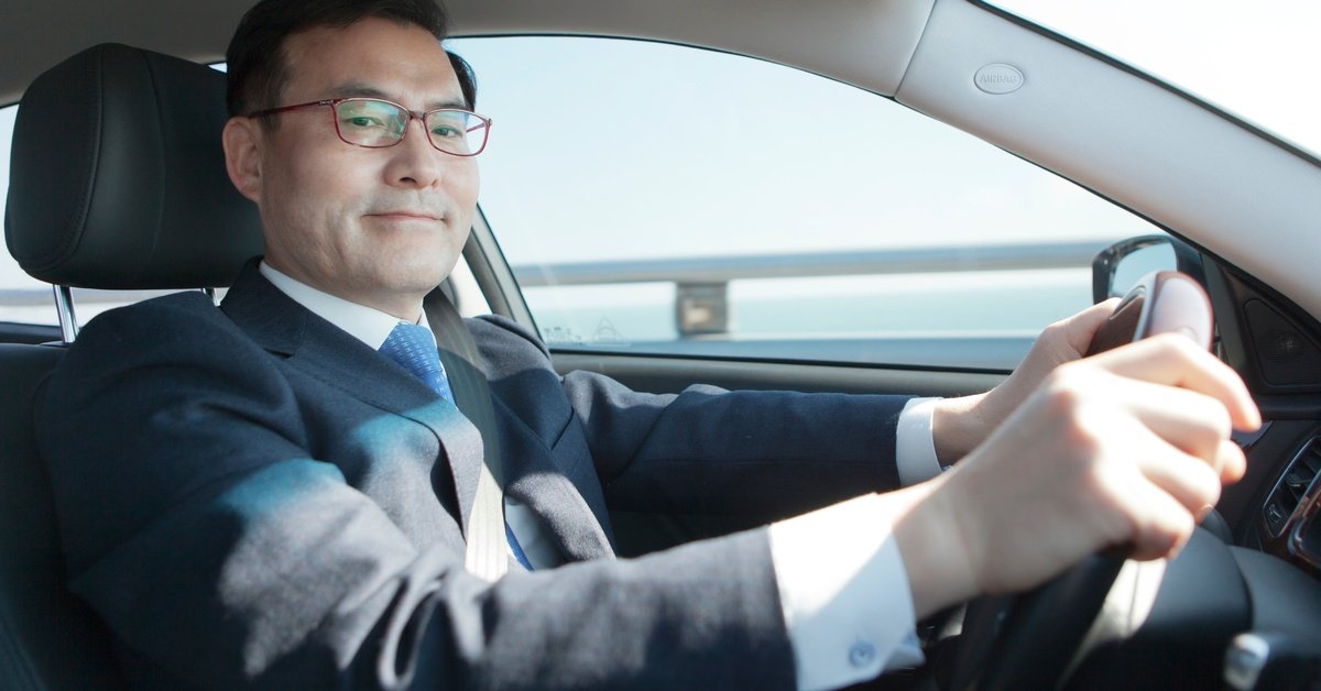 A man wearing a business suit and glasses smiles as he drives a car. His driver's side window offers a view of a lake.