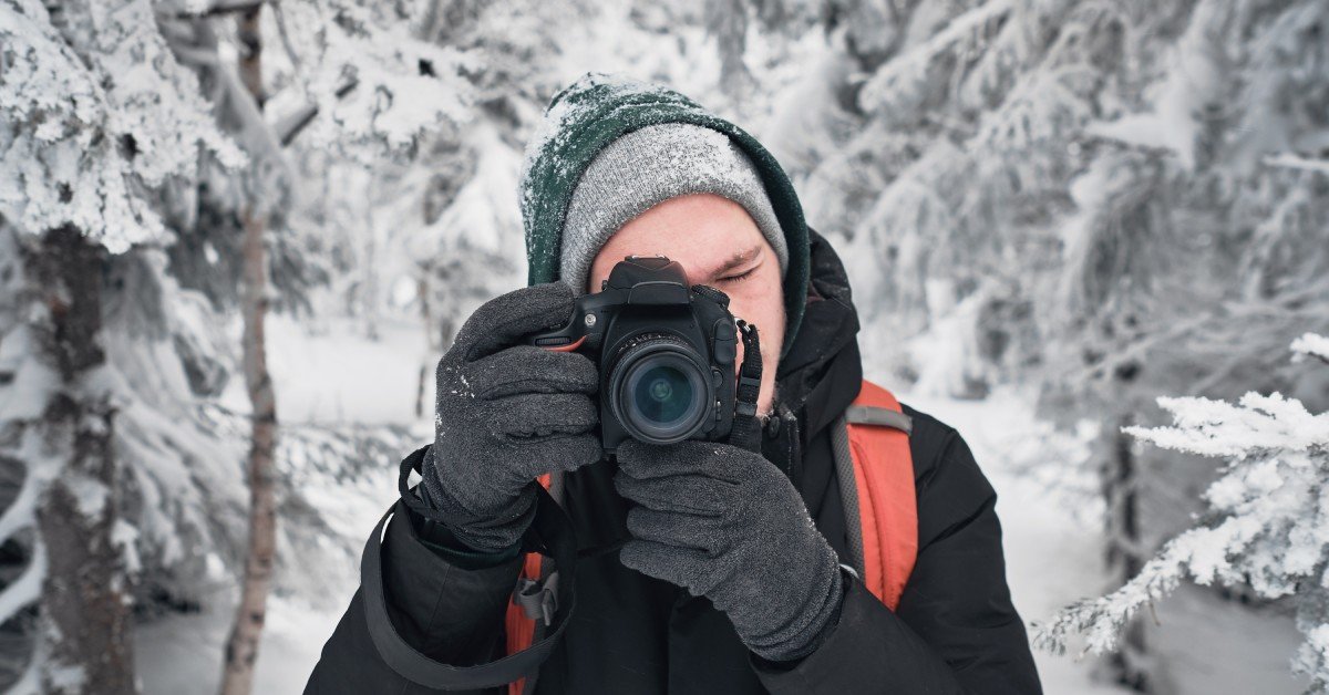 A person wearing a coat, hat, and gloves holds a camera to his eye. Snow covers the ground and trees in the background.