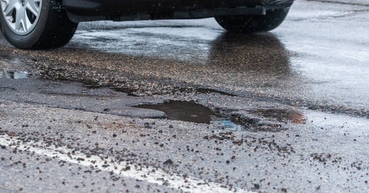 A wet, rainy road with a massive pothole filled with rainwater. There is a car approaching in the background.