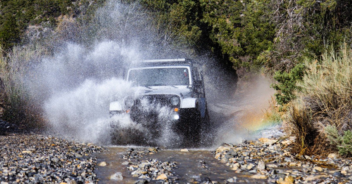 A white off-road vehicle with a roof rack splashes through a rocky stream, creating a dramatic water spray and a rainbow effect.