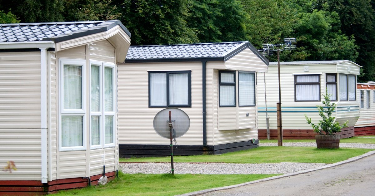 Three mobile homes sit next to each other in a trailer park. One has a satellite dish on its lawn, and another has a tree.