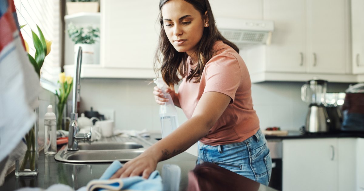 A woman stands in her kitchen and cleans her counters. She holds a spray bottle and a soft blue cloth.