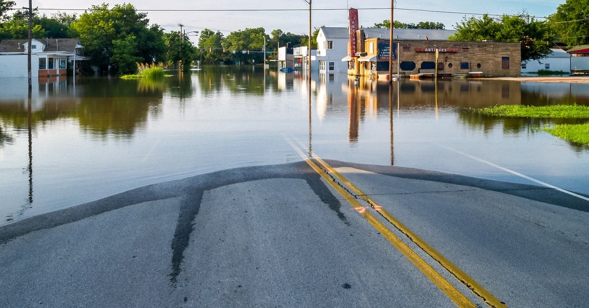 A flooded street with submerged buildings, utility poles, and trees, reflecting in calm water under a partly cloudy sky.