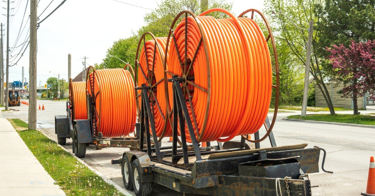 Two trailers parked parallel on the street curb with orange spools of brand-new fiber conduit and orange cones behind them.