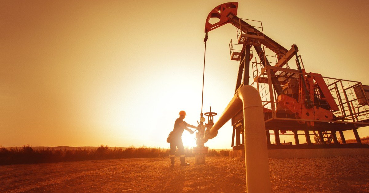 An oil and gas industry worker stands next to the oil rig as the sun sets behind them. They wear a hard hat and uniform.