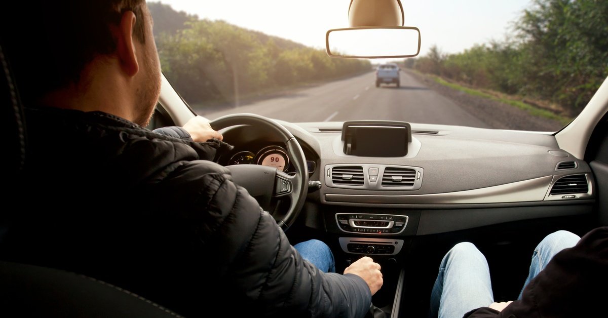 A man is sitting in the driver’s seat of a car as he drives down a road. There is a truck ahead of him.