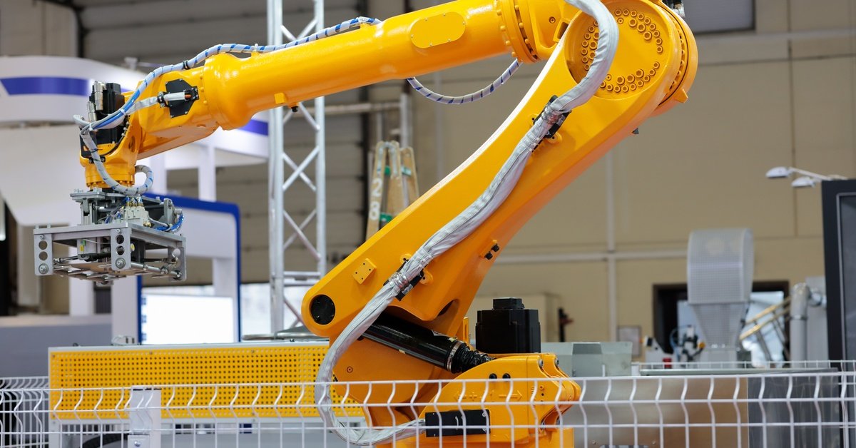 A bright yellow industrial robotic arm with a gripper is in a factory, partially behind a white safety fence.