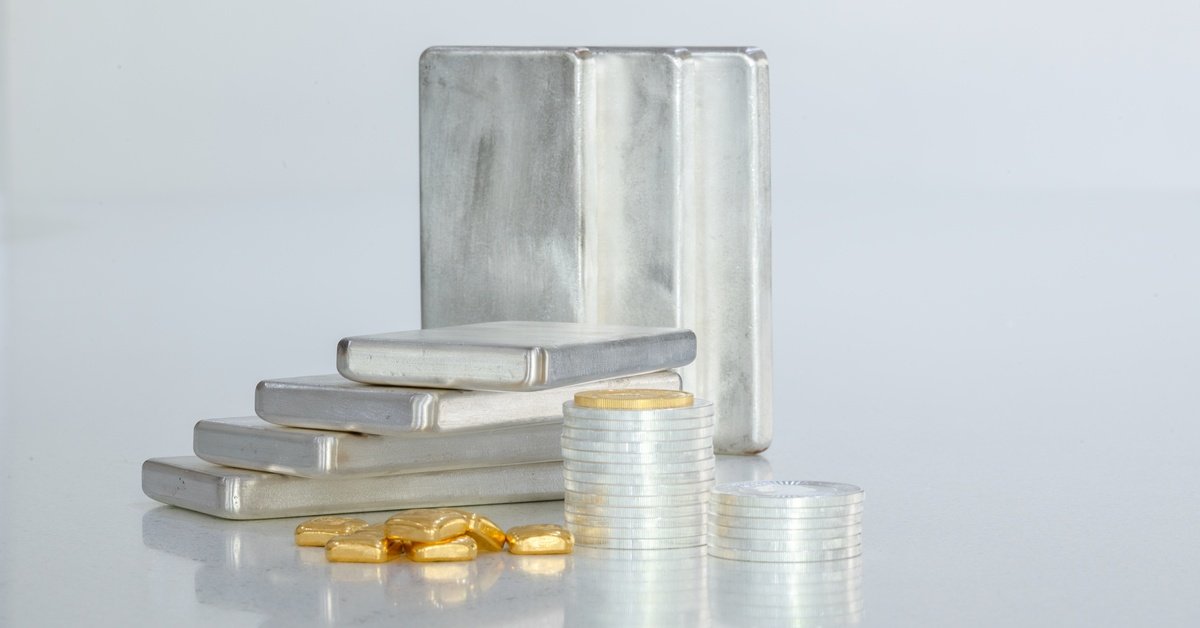 Stacks of silver coins sit in front of a stack of silver bars and a pile of gold bars. The background is white.