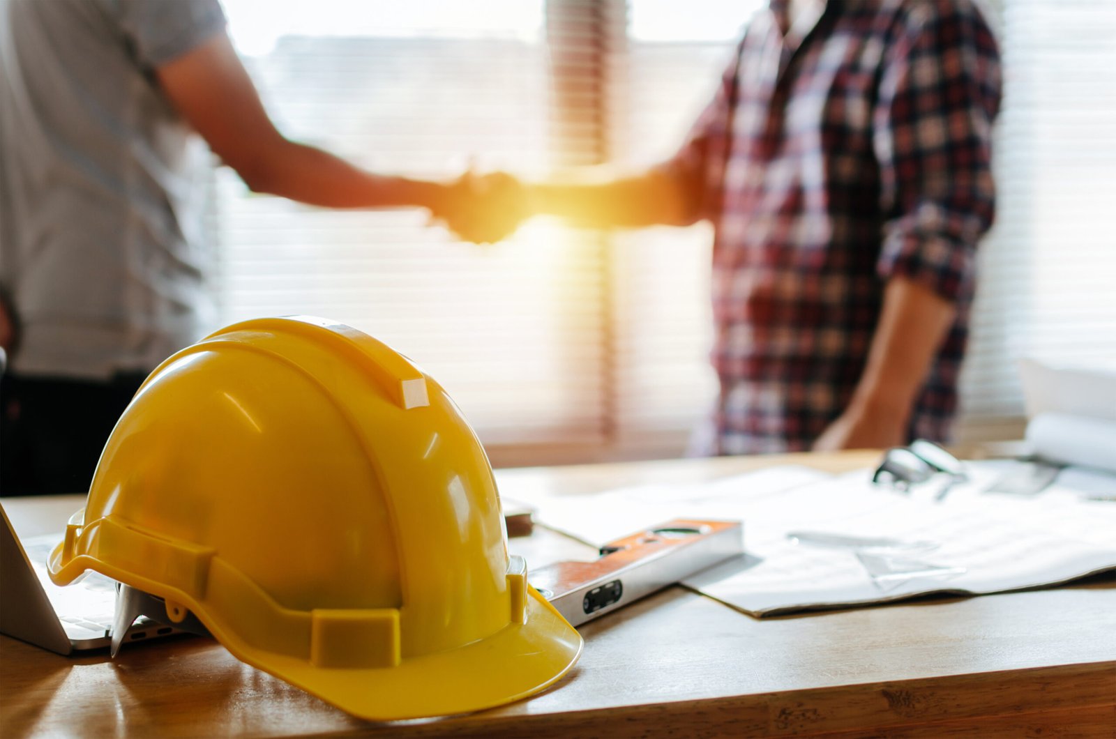 Two men shaking hands behind a desk covered in blueprints and construction gear, including a hard hat and level.
