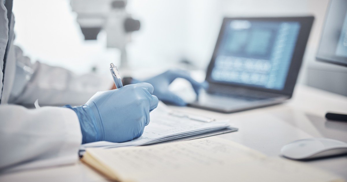 A gloved researcher writes notes on a clipboard at a lab desk, with a laptop and a microscope blurred in the background.