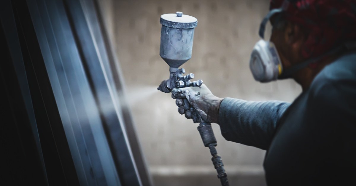 A worker wearing a respirator and gloves uses a spray gun to coat a metal panel in an industrial workshop.