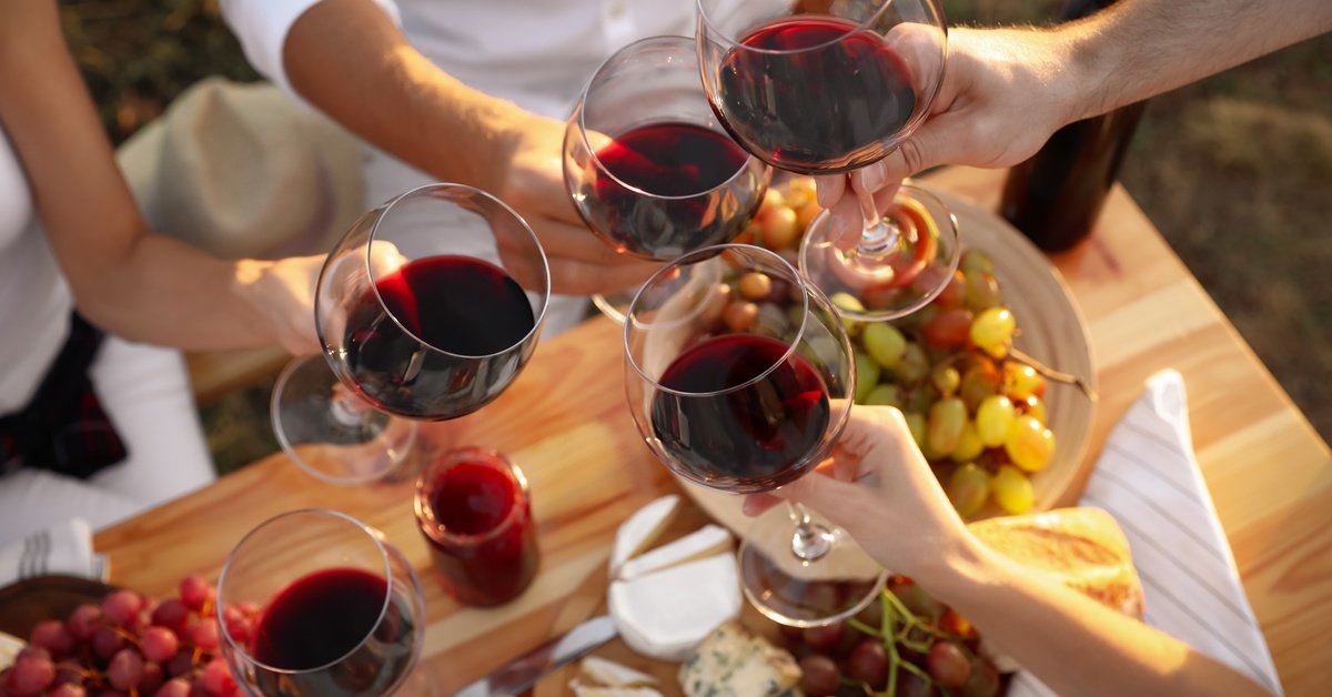 A group of people clink glasses of red wine over a wooden table with grapes, cheese, and sliced bread in warm sunlight.