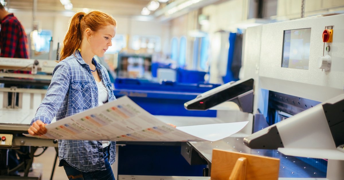 A woman wearing a blue plaid long-sleeve shirt is holding a large print next to an industrial printer in a print shop.