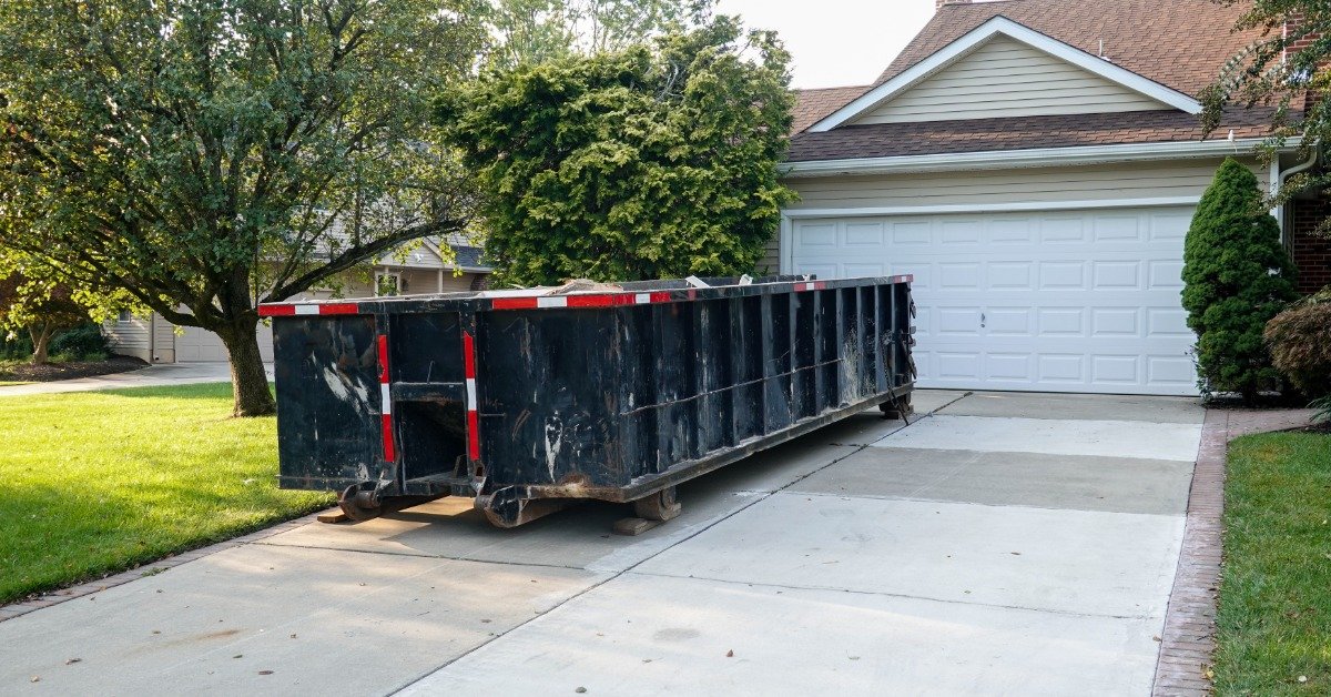 A large black construction dumpster on a suburban driveway in front of a garage, with trees and a house in the background.