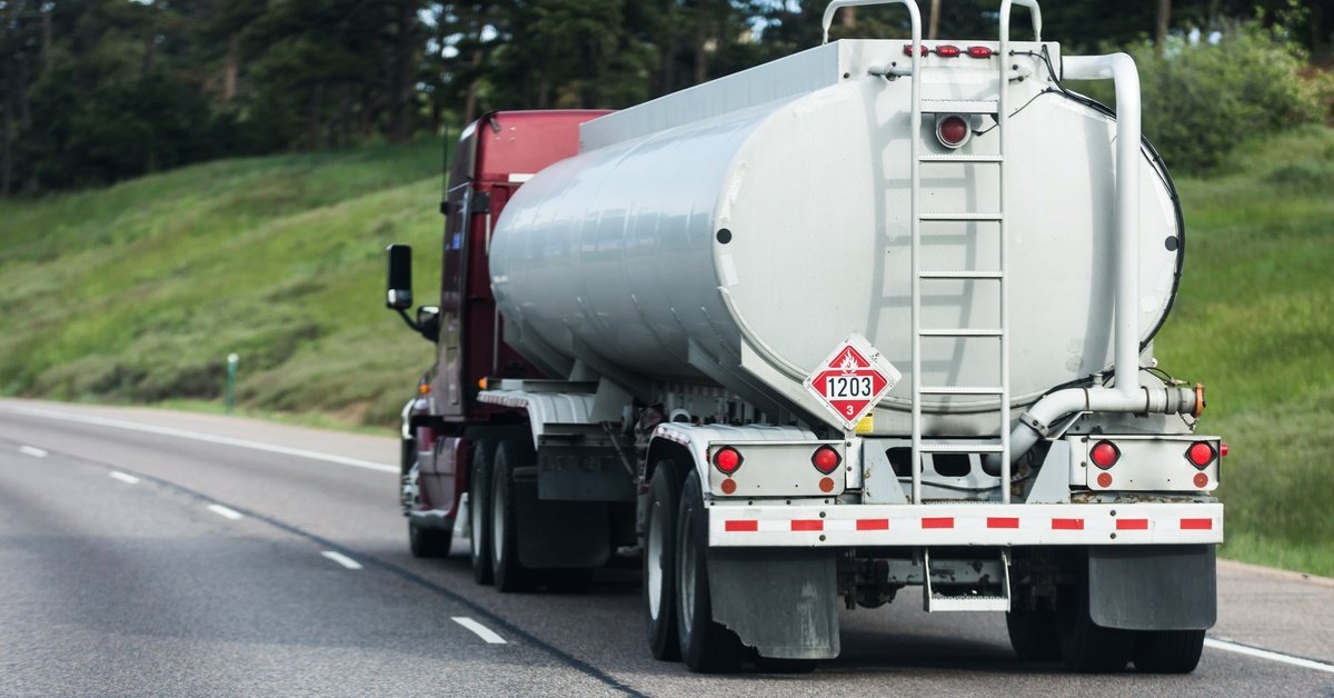A rear view of a truck with a large white cylindrical tank driving on a highway, surrounded by green trees.