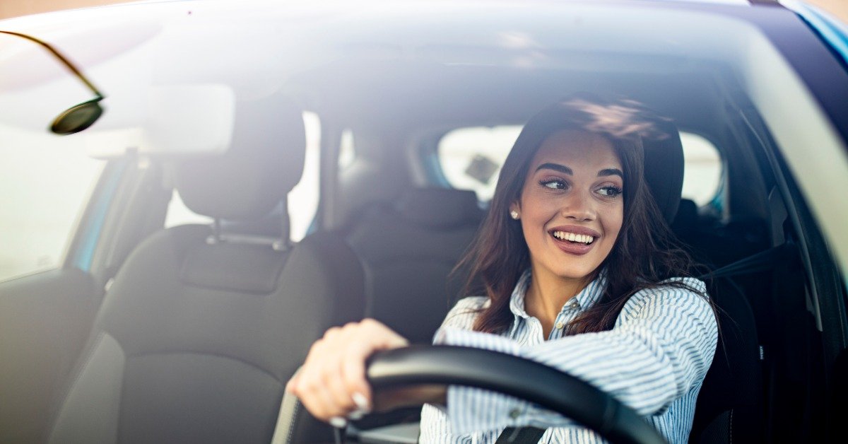 A smiling woman driving a car with her hands on the steering wheel while sunlight streams through the windshield.