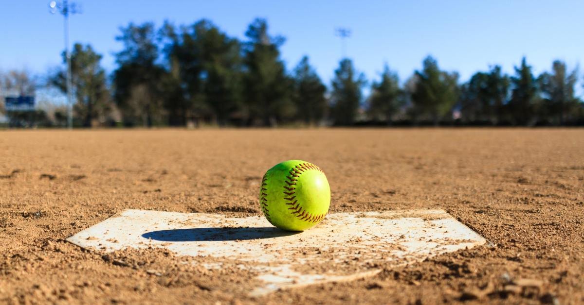 A close-up view shows a bright-yellow softball sitting on a home plate that's covered with scattered sand.
