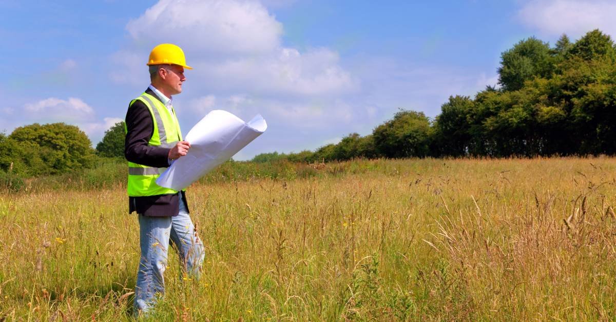 A land architect wears a yellow hard hat in the field as they hold a map of the land to design a functional space.
