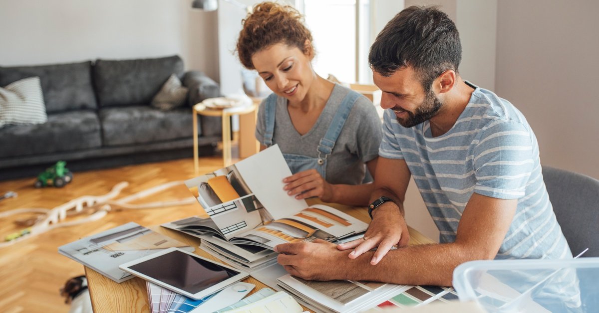 A couple sitting at a table with furniture catalogs and paint samples for a home renovation. A tablet is on the table.